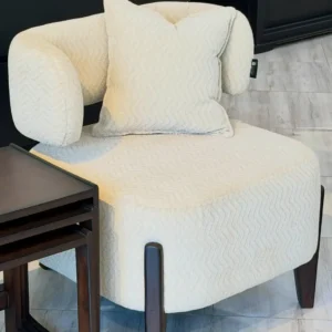 Cream accent chair Kenya with rounded arms, textured upholstery, matching pillow, and dark legs