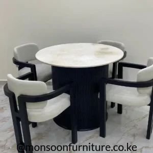 Round dining table with pale top, black ribbed pedestal base and four cream upholstered chairs with black frames.