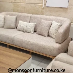 Beige textured sofa with continuous rounded back, broad seat cushions, neutral pillows and a slim warm wood-tone base.