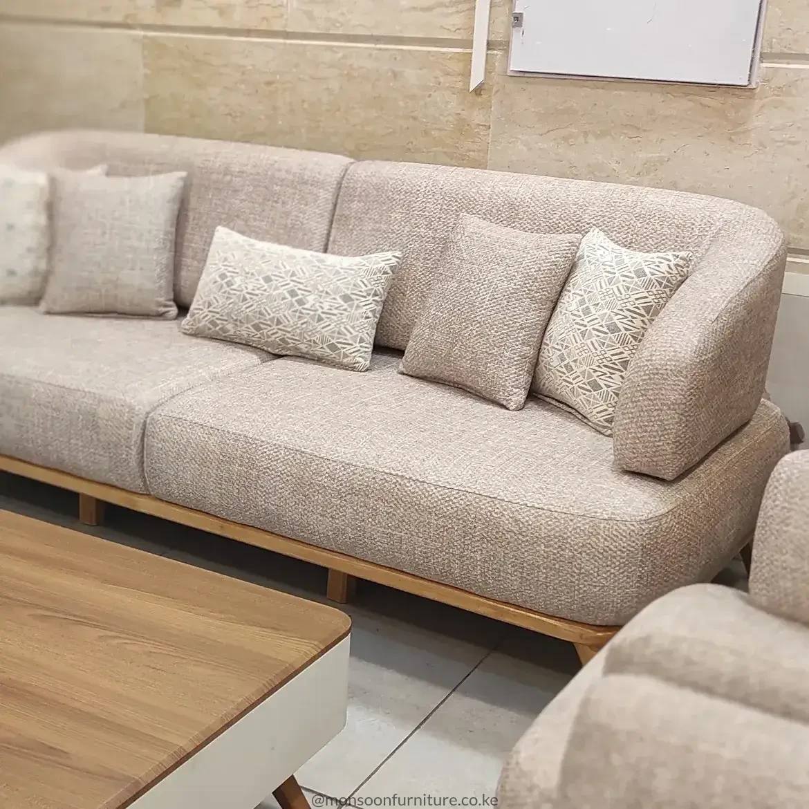 Beige textured sofa with continuous rounded back, broad seat cushions, neutral pillows and a slim warm wood-tone base.