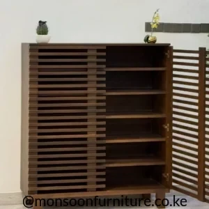 Dark brown shoe cabinet with horizontal slats, a staggered centre pattern, flat top and raised legs against a light wall.