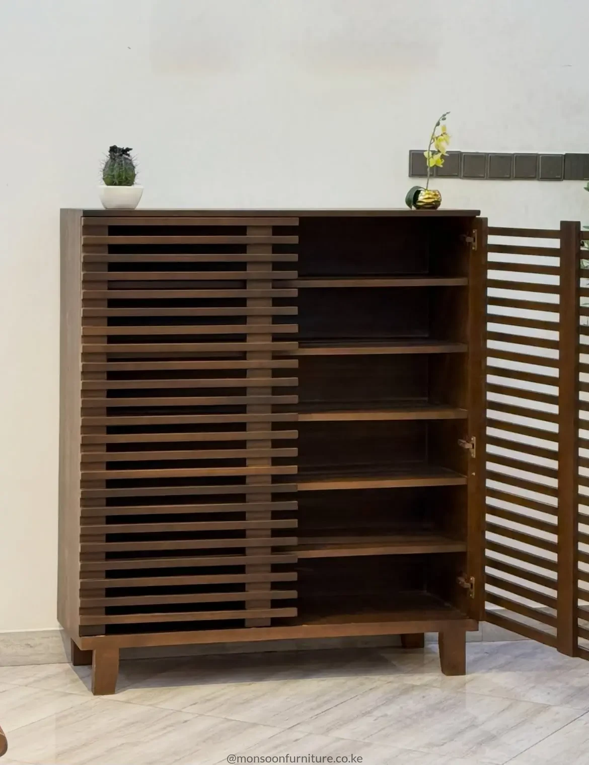 Dark brown shoe cabinet with horizontal slats, a staggered centre pattern, flat top and raised legs against a light wall.
