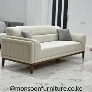 Cream sofa with vertical channel detailing, rounded arms, warm brown raised base and two grey cushions on marble-look flooring.