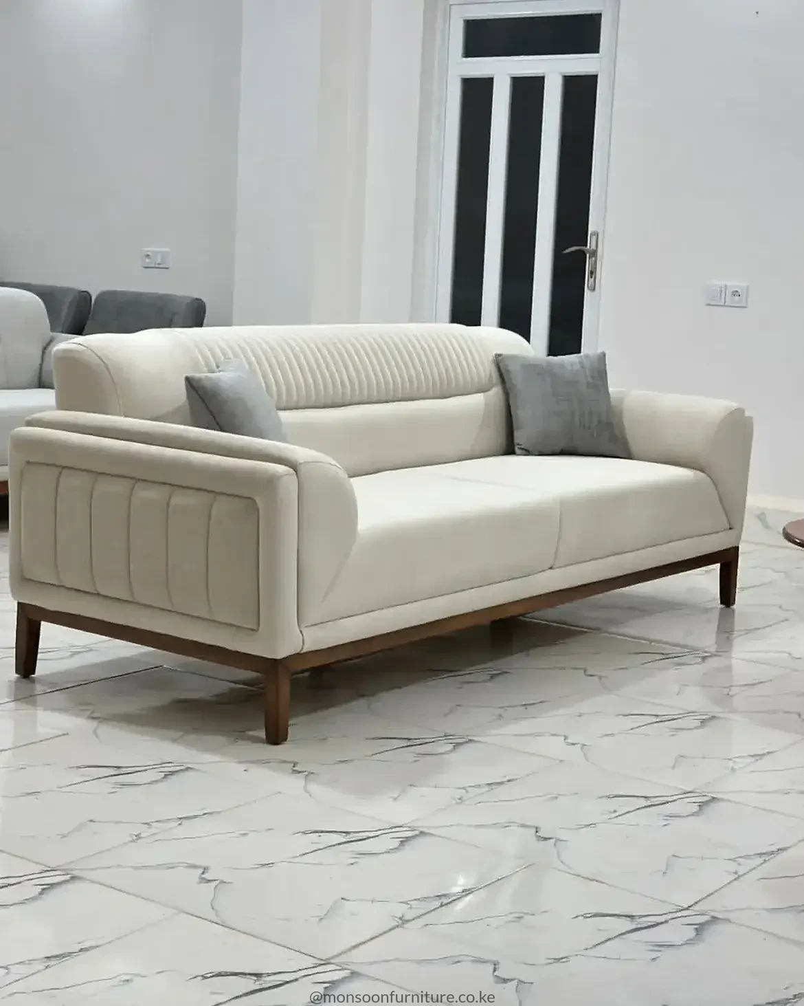 Cream sofa with vertical channel detailing, rounded arms, warm brown raised base and two grey cushions on marble-look flooring.