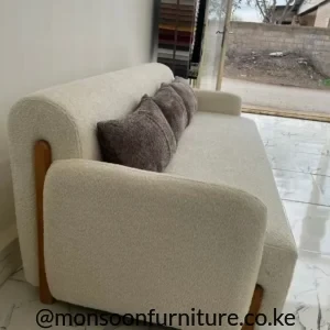 Cream textured sofa with rounded back and arms, warm brown-toned side accents and three brown-grey cushions in a bright showroom