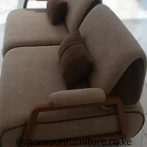 Taupe textured sofa with rounded back cushions, angular warm brown side frame, padded armrest and darker brown accent cushions.