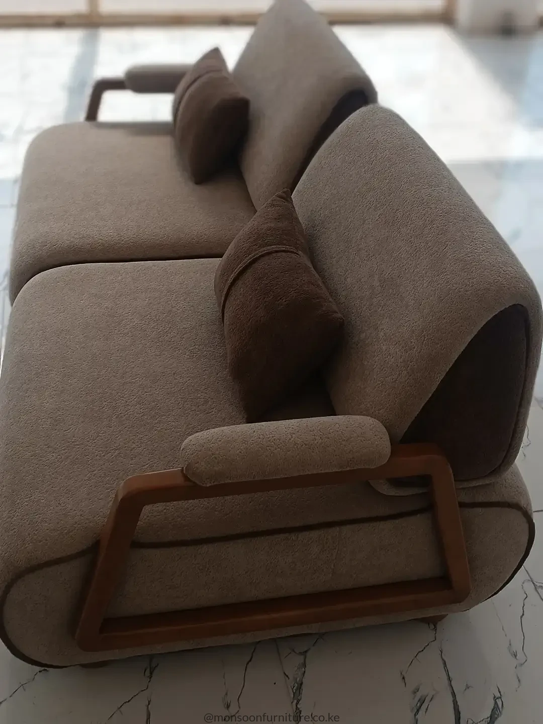 Taupe textured sofa with rounded back cushions, angular warm brown side frame, padded armrest and darker brown accent cushions.