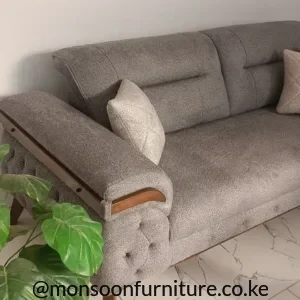 Warm grey textured sofa with button-detailed outer arm, brown frame-style trim, tapered legs and a light accent cushion.