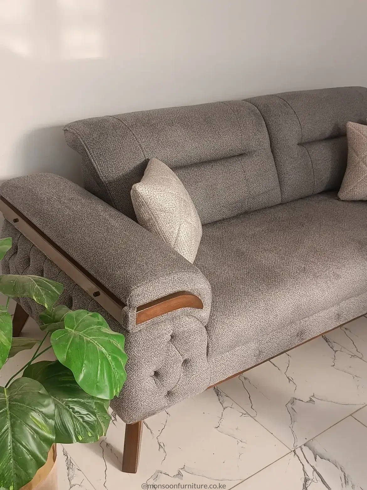Warm grey textured sofa with button-detailed outer arm, brown frame-style trim, tapered legs and a light accent cushion.