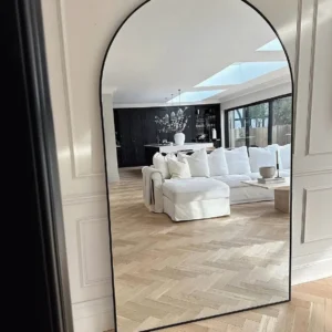 Black arched full size mirror with slim dark frame leaning against a panelled wall in a bright living room