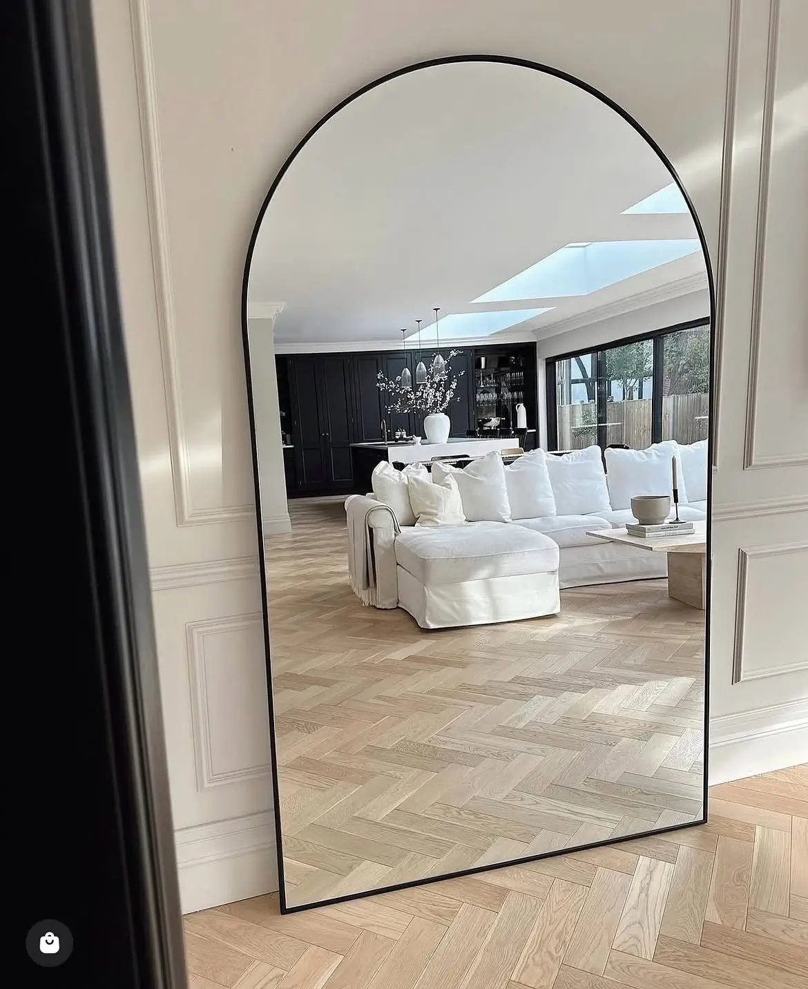 Black arched full size mirror with slim dark frame leaning against a panelled wall in a bright living room