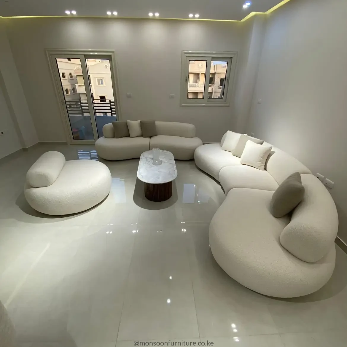 Cream curved upholstered seating with rounded low-profile forms, neutral cushions and a central oval coffee table in a bright living room.
