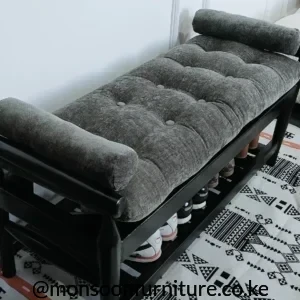 Grey tufted cushioned shoe storage bench with a dark frame, cylindrical bolsters and open lower shelving holding shoes.