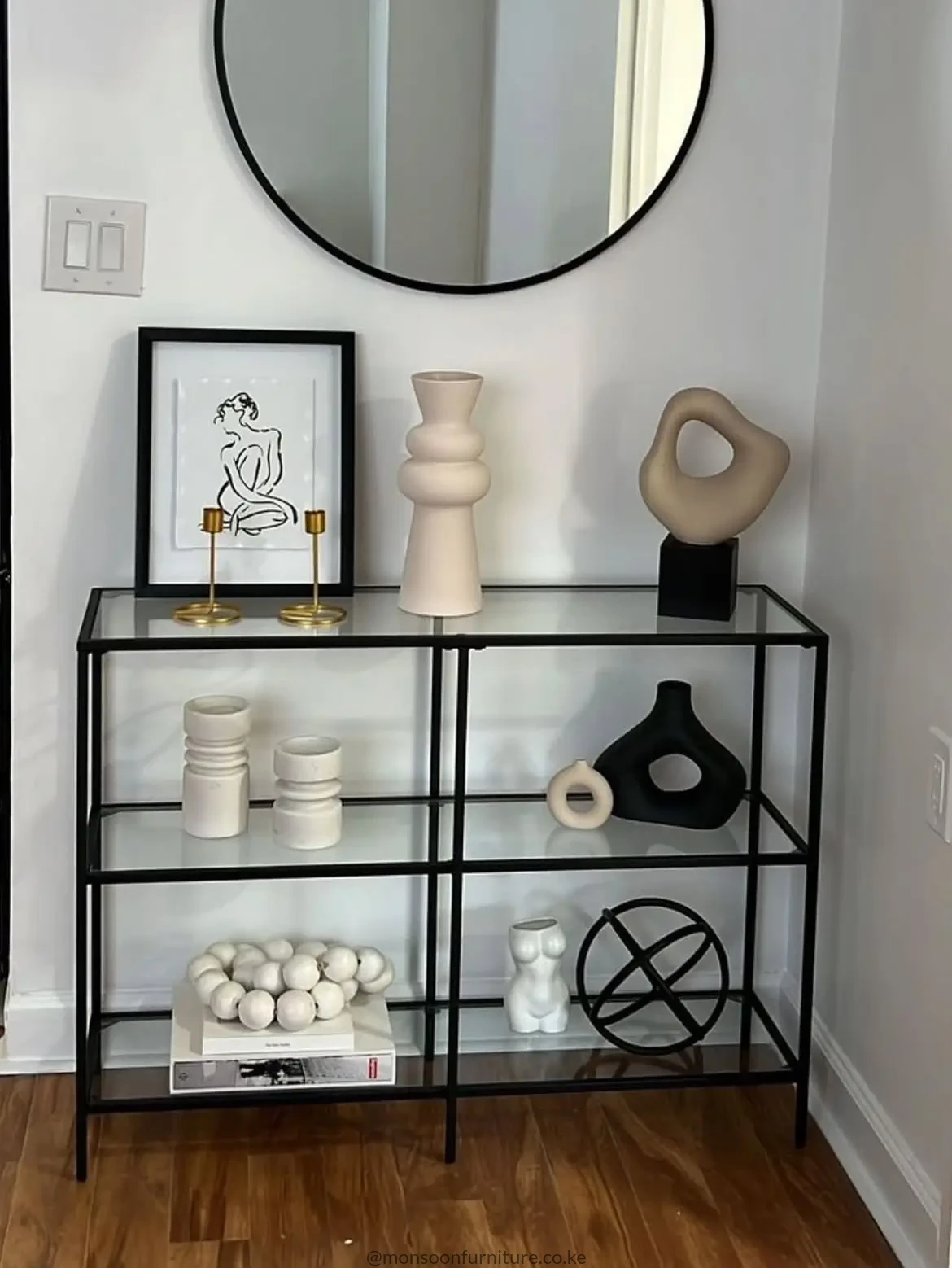Black small console table with clear glass shelves and round black-framed mirror in a bright hallway