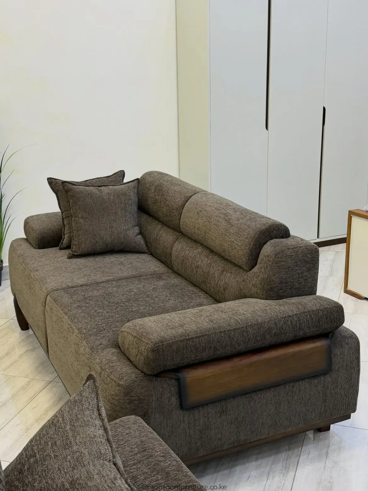 Taupe-brown sofa with textured upholstery, broad padded arms, segmented back cushions and wood-tone side accents in a bright room.