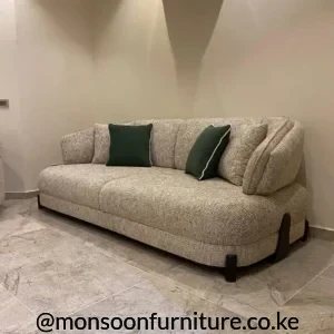 Taupe curved sofa with textured upholstery, rounded arms, dark green cushions and short dark feet on a grey tiled floor.