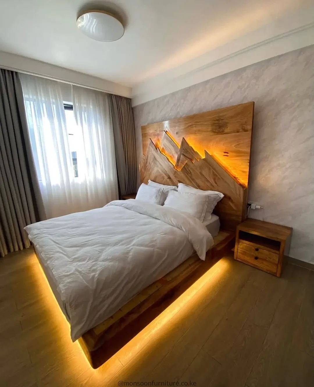 Warm brown wooden platform bed with angular illuminated headboard and warm under-bed lighting beside a two-drawer bedside table.